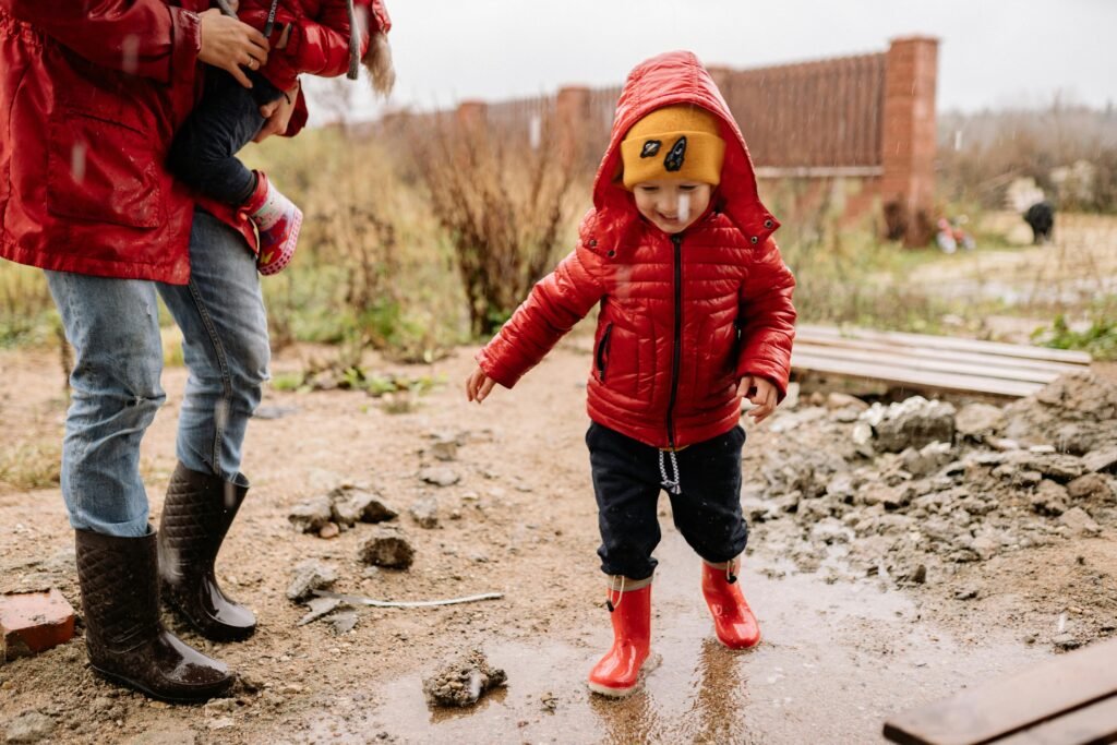 A young child in a red jacket and beanie joyfully splashes in a puddle.